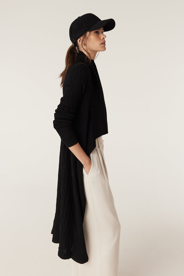 Woman wearing a black merino wool longline cable knit cardigan, black cap, and cream pants.