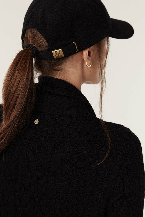 Woman wearing a black Merino wool cable knit cardigan with a ribbed collar and a black baseball cap.