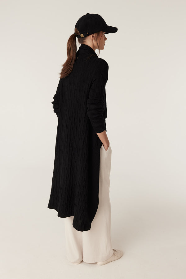 Black Merino wool longline cardigan with cable knit detail, worn by a model with a black cap.