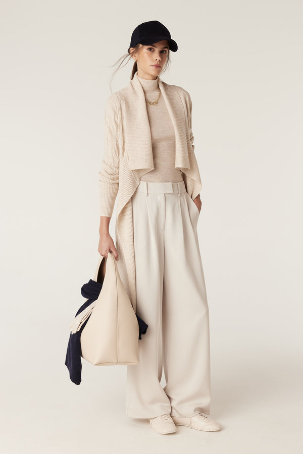 Model wears an oatmeal Merino wool longline cable knit cardigan, cream wide-leg pants, and a black cap.