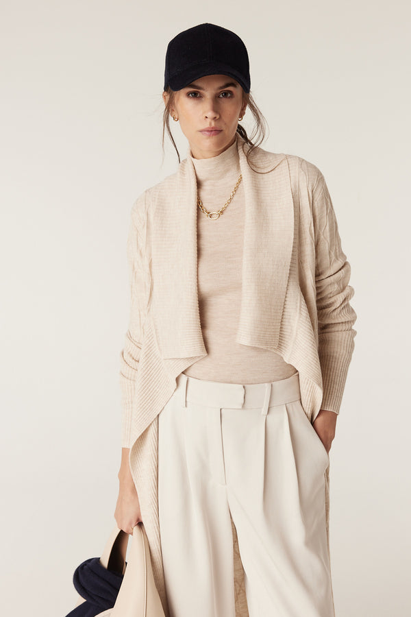 Woman in an oatmeal merino wool cable knit longline cardigan, cream turtleneck, and pants, wearing a black cap.