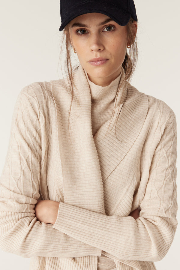 Woman in an oatmeal merino wool cable knit cardigan with a ribbed collar, arms crossed.