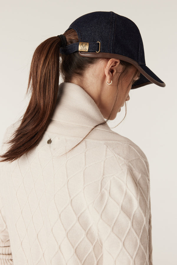 Woman in antique white Merino wool cardigan with diamond cable stitch and a dark denim baseball cap.