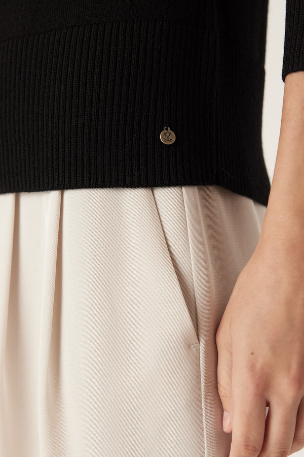 Black Merino wool crew neck jumper with ribbed hem and gold branded plate, worn with light trousers.