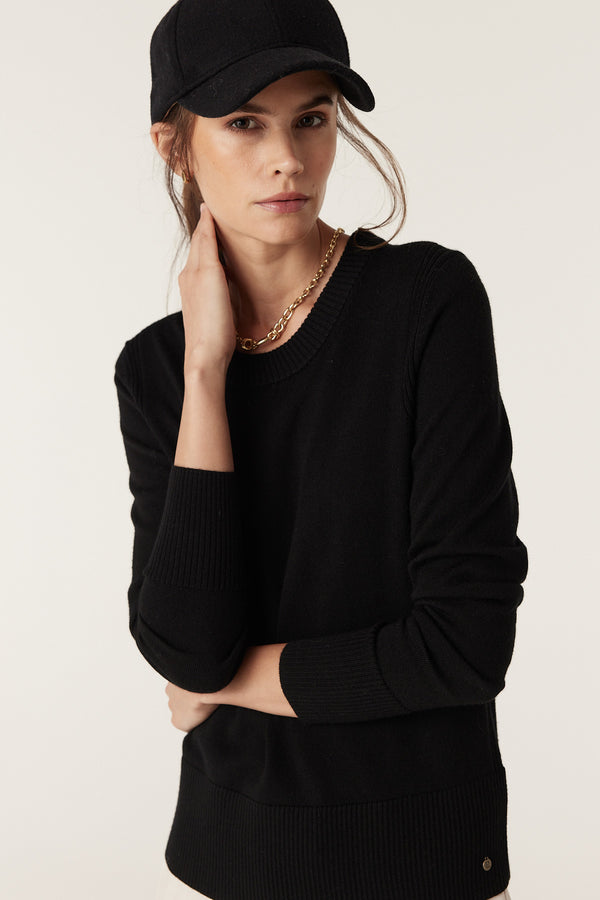 Woman in a black merino wool crew neck jumper with rib trims, wearing a black cap and gold chain.