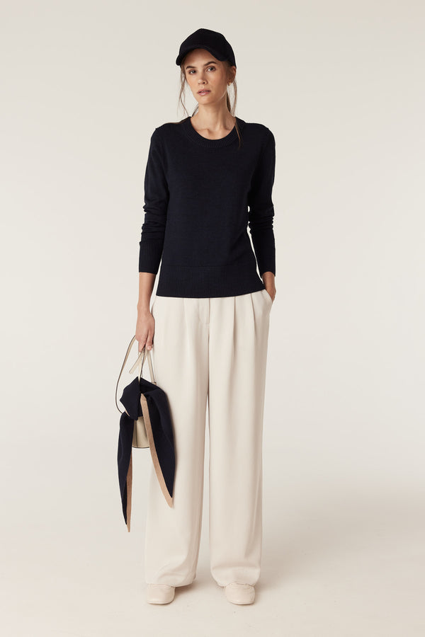 Woman in a midnight navy Merino wool crew neck jumper, cream wide-leg trousers, and a black cap.