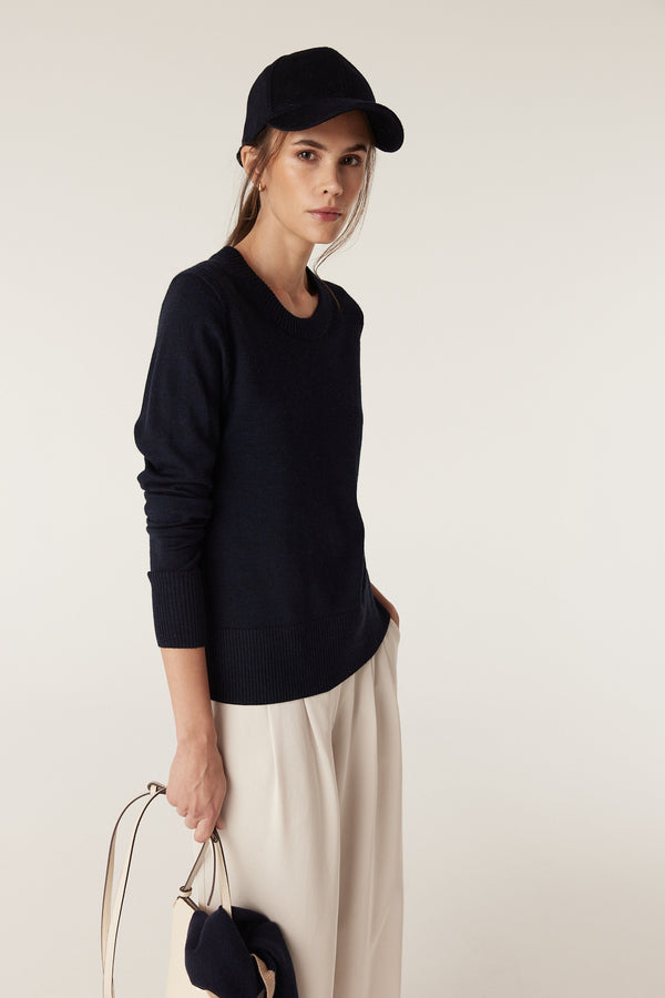 Woman in a midnight navy Merino wool crew neck jumper, light trousers, a navy cap, and holding a bag.