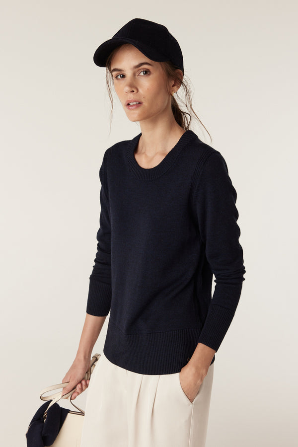 Woman in a Midnight Navy Merino wool crew neck jumper with rib trims, light pants, and a navy cap.