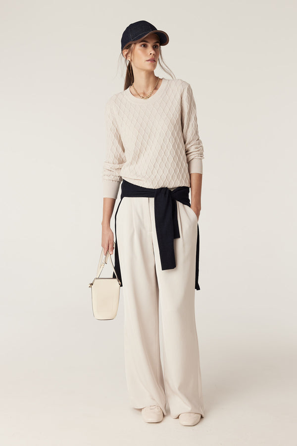 Woman in antique white merino wool jumper with diamond cable stitch, cream pants, and navy sweater tied at waist.