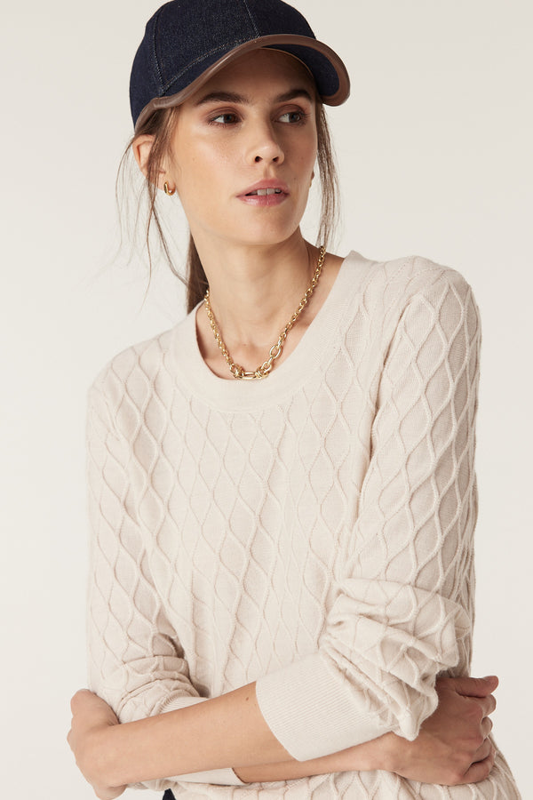 Woman in antique white merino wool jumper with diamond cable stitch, wearing a baseball cap and gold necklace.