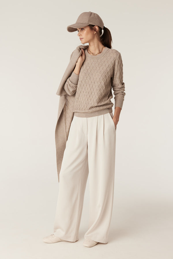 Woman in pebble merino wool jumper with diamond cable stitch, cream pants, and matching cap.
