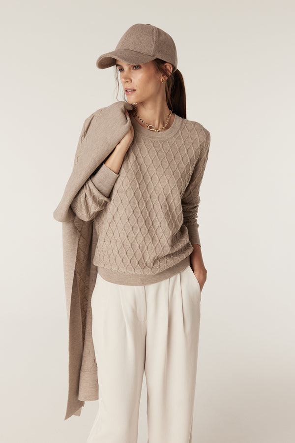 Woman in pebble merino wool jumper with diamond cable stitch, cream pants, and matching cap.
