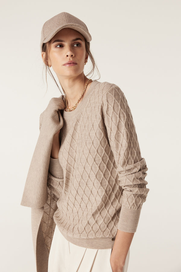 Woman in pebble merino wool jumper with diamond cable stitch, matching cap, and gold chain.