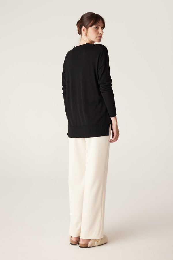 Model in black merino wool V-neck jumper with dropped shoulders and high-low hem, paired with cream pants.