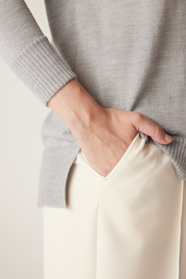 Grey marle merino wool jumper with ribbed cuffs, side split hem, and hand in cream pants pocket.