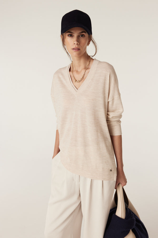 Model wears an oatmeal Saxon Fine Merino Jumper with V-neck, dropped shoulders, and high-low hem.