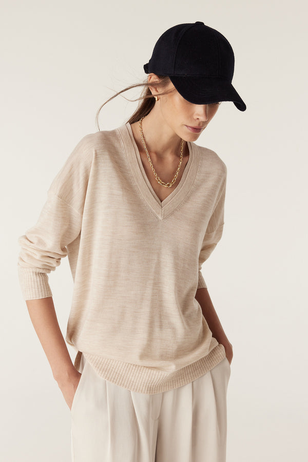 Woman in oatmeal fine merino wool V-neck jumper with dropped shoulders, wearing a black cap and gold chain.