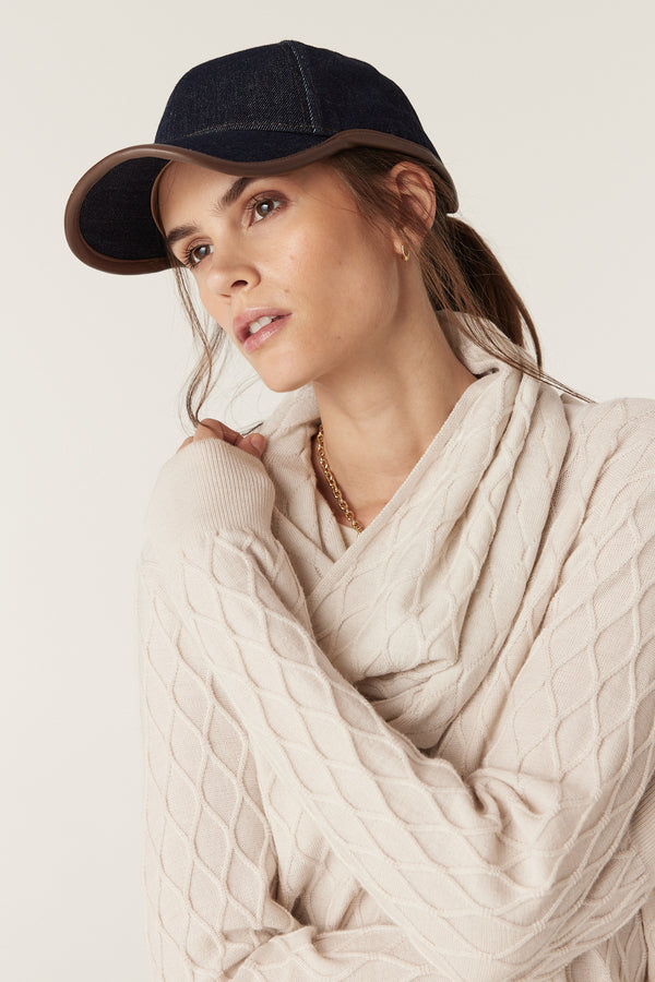 Woman wearing an antique white merino wool snood with diamond cable stitching and a dark denim baseball cap.
