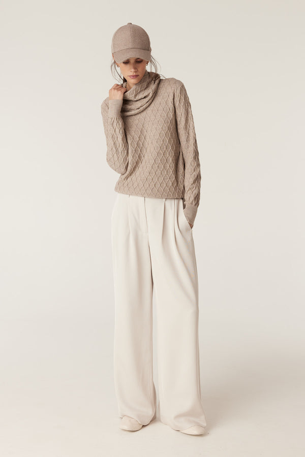 Model wearing a pebble merino wool snood with diamond cable stitching, light pants, and a matching cap.