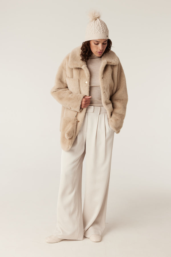 Woman in an oatmeal merino beanie with a faux fur pom pom, paired with a beige faux fur jacket and cream pants.