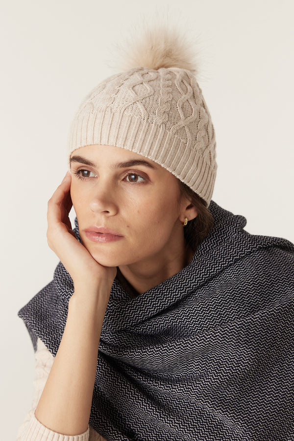 Oatmeal merino wool beanie with diamond cable knit, ribbed hem, and faux fur pom pom on a model.