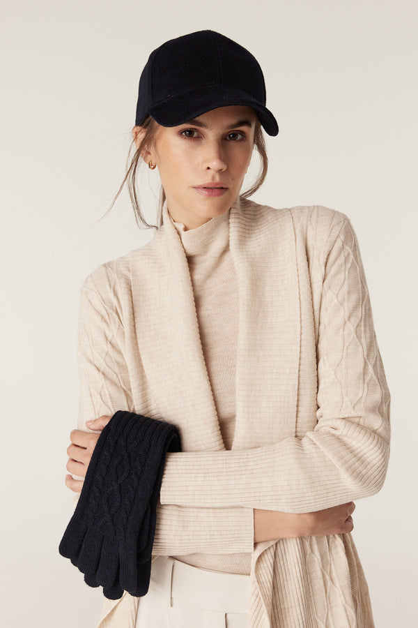 Woman wearing a cream merino cardigan and turtleneck, holding navy merino cable knit gloves.