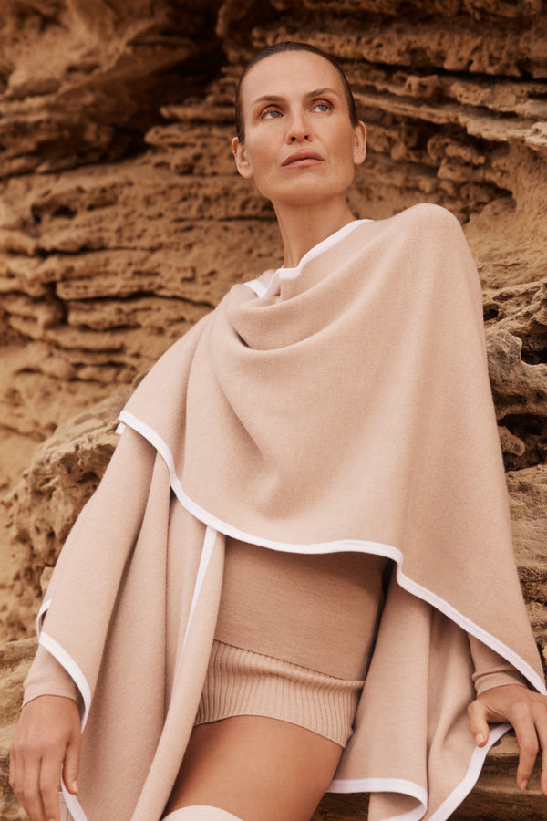 Latte Glenelg Merino Cape with white contrast trim, worn by a woman against a textured rock wall.