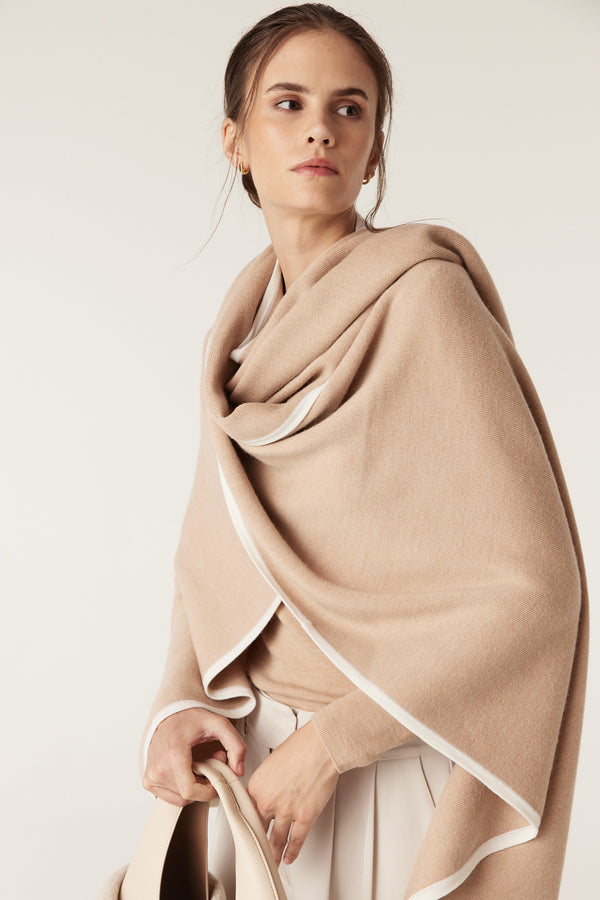 Model wearing a latte merino wool cape with white contrast trim, draped elegantly over a light top.