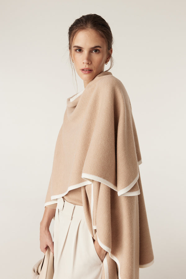 Woman wearing a latte merino wool cape with white contrast trim, layered over light-colored clothing.