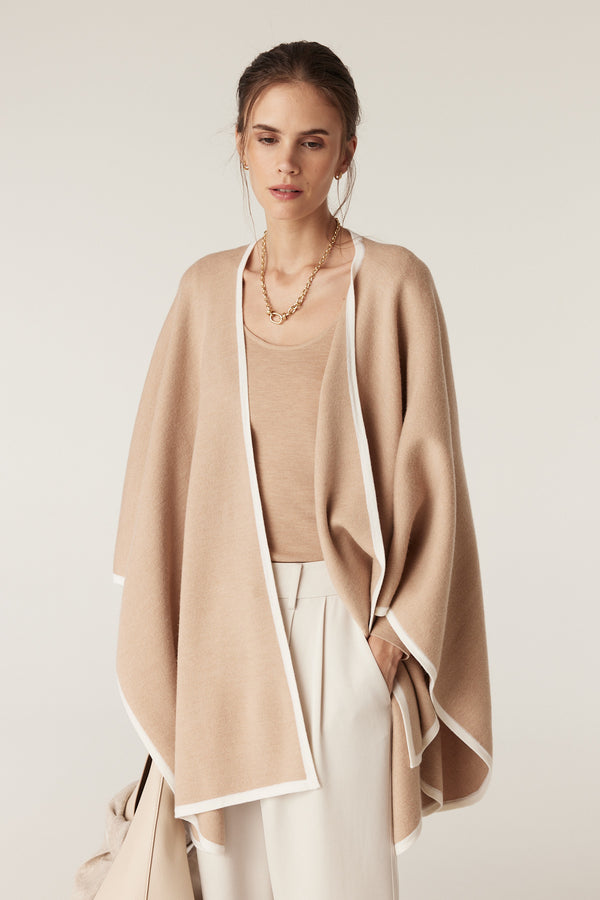 Model wearing a latte merino wool cape with white contrast trim over a matching top and white pants.