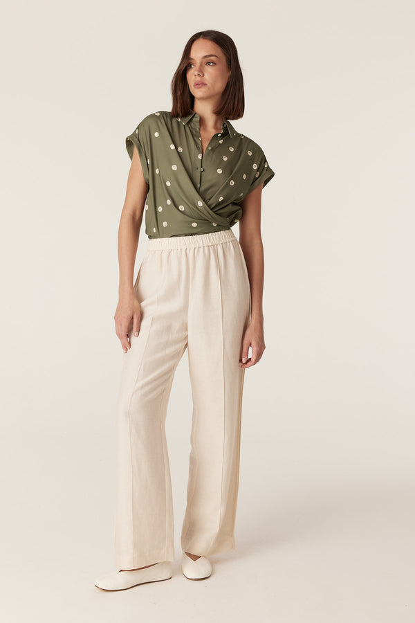 Olive green collared blouse with white polka dots, featuring a wrap-front design and cap sleeves, paired with cream pants.