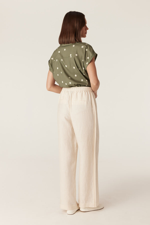 Woman wearing a green collared blouse with white polka dots, cap sleeves, and cream-colored wide-leg trousers.