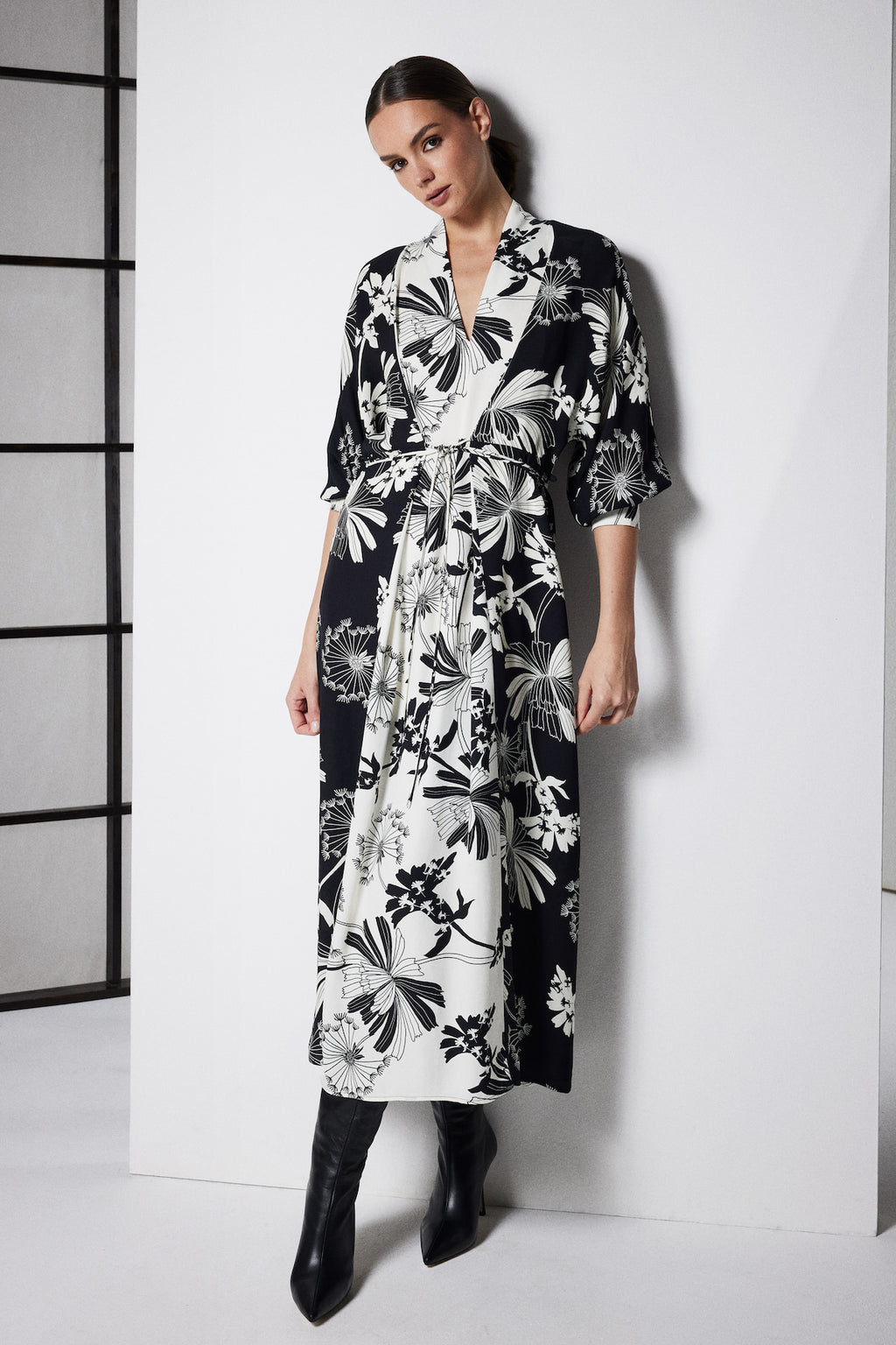 Model wearing a black and white deco print midi dress with a V-neck, batwing sleeves, and a wraparound waist tie.
